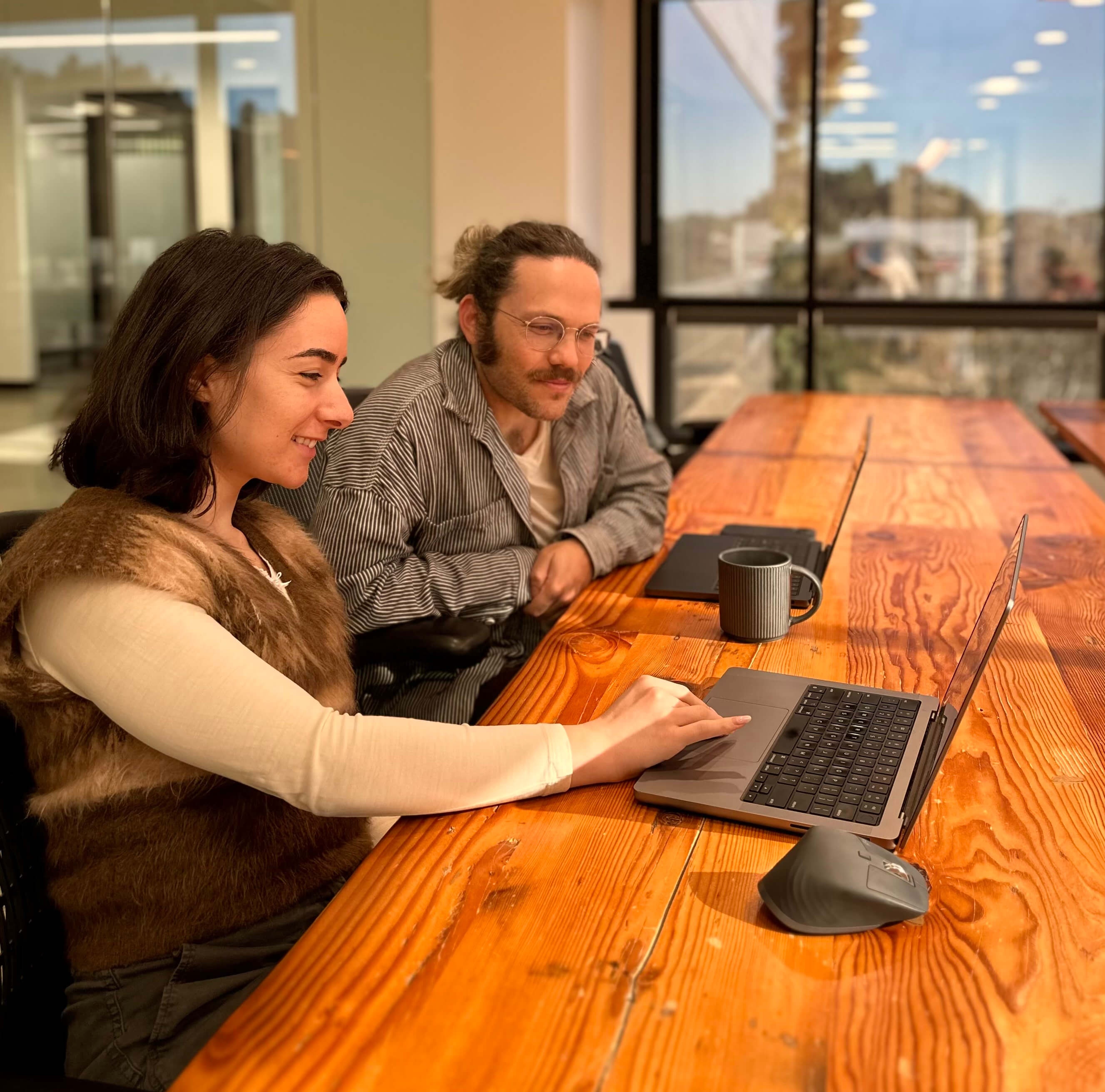 Two people sitting at a wooden table collaborating while looking at a laptop screen.